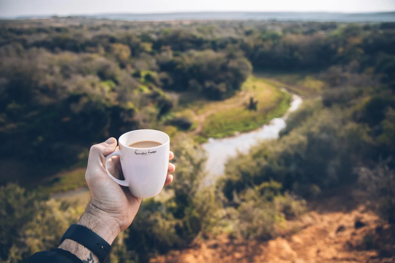 A hand holding a white mug above a wooded valley and winding watercourse